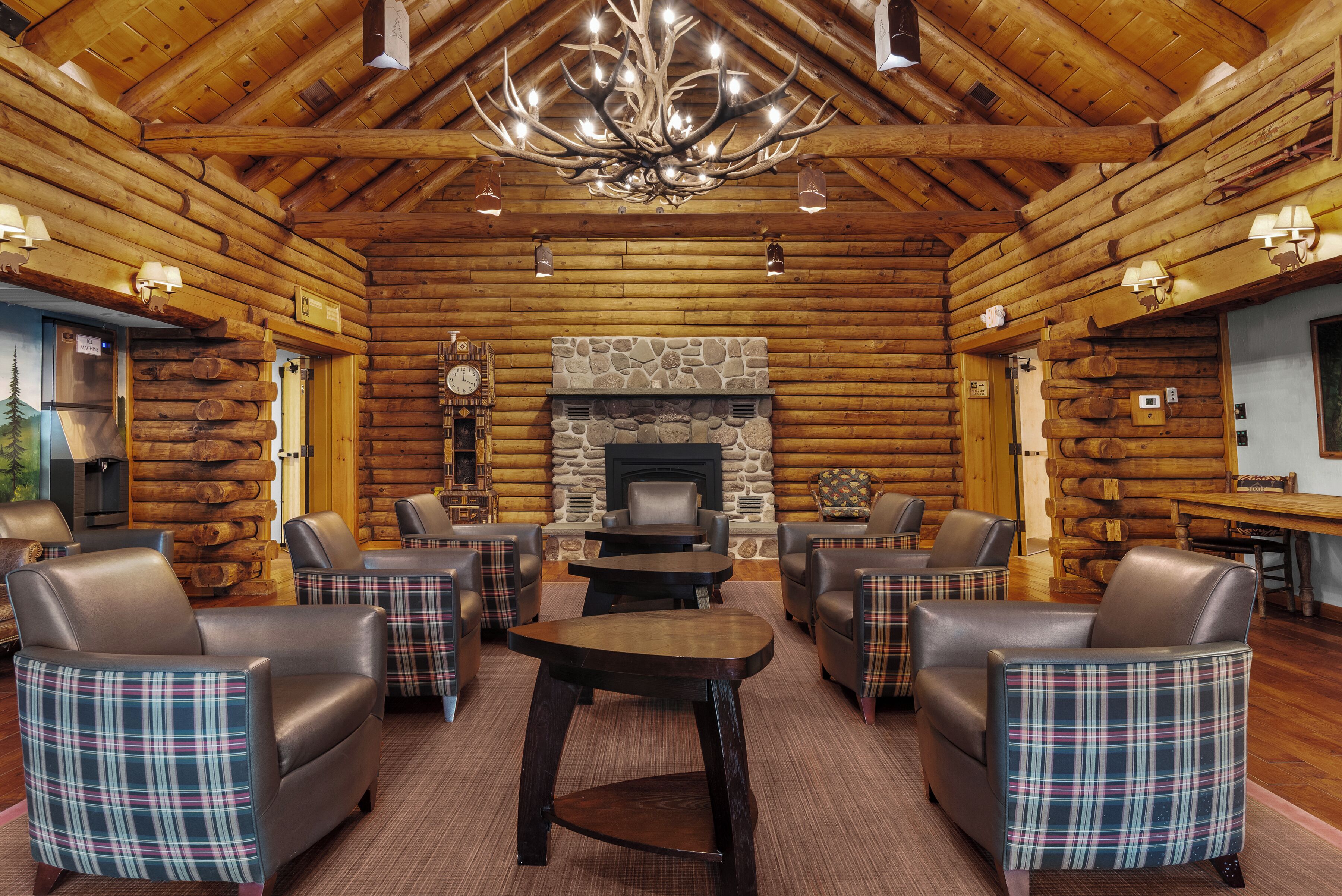 A log cabin interior with a stone fireplace, leather chairs, a wooden table, and a chandelier.