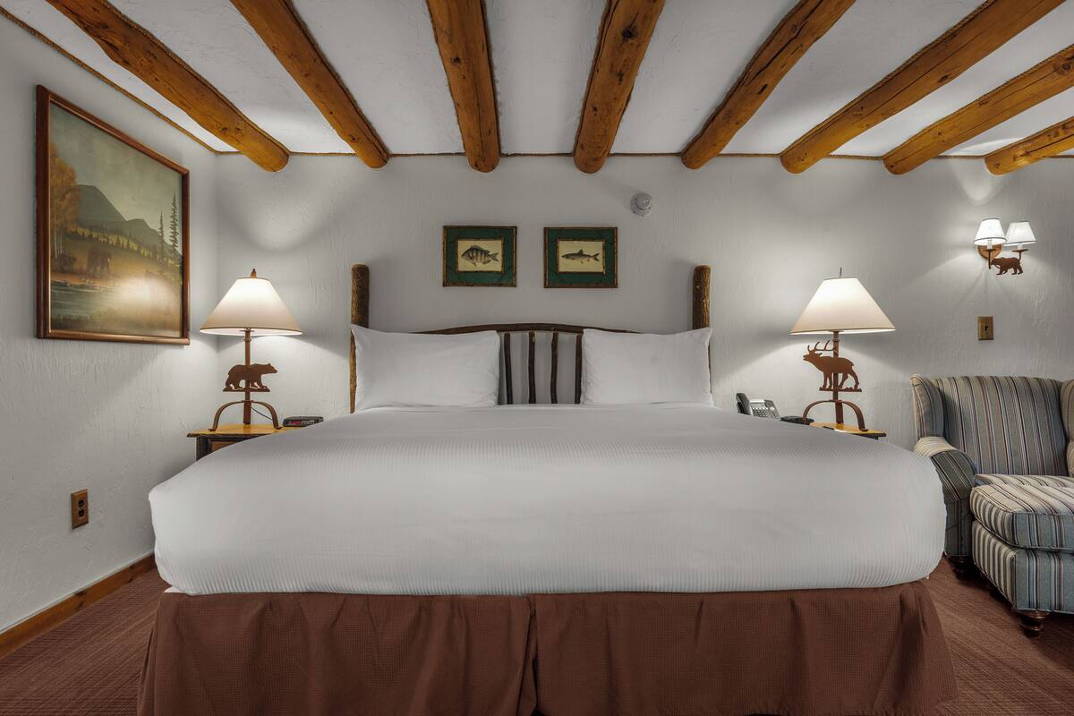 A large bed with a wooden headboard, two bedside lamps, a chair, and two framed pictures on the wall.