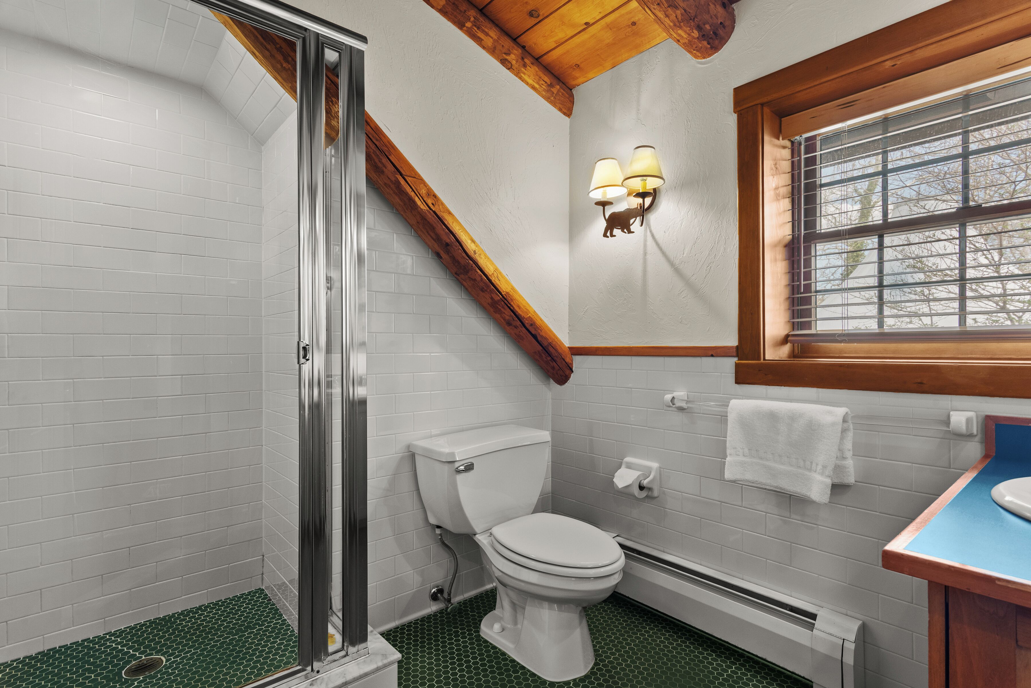 A bathroom with a shower, toilet, and sink, featuring white tiles, a wooden ceiling, and a window.