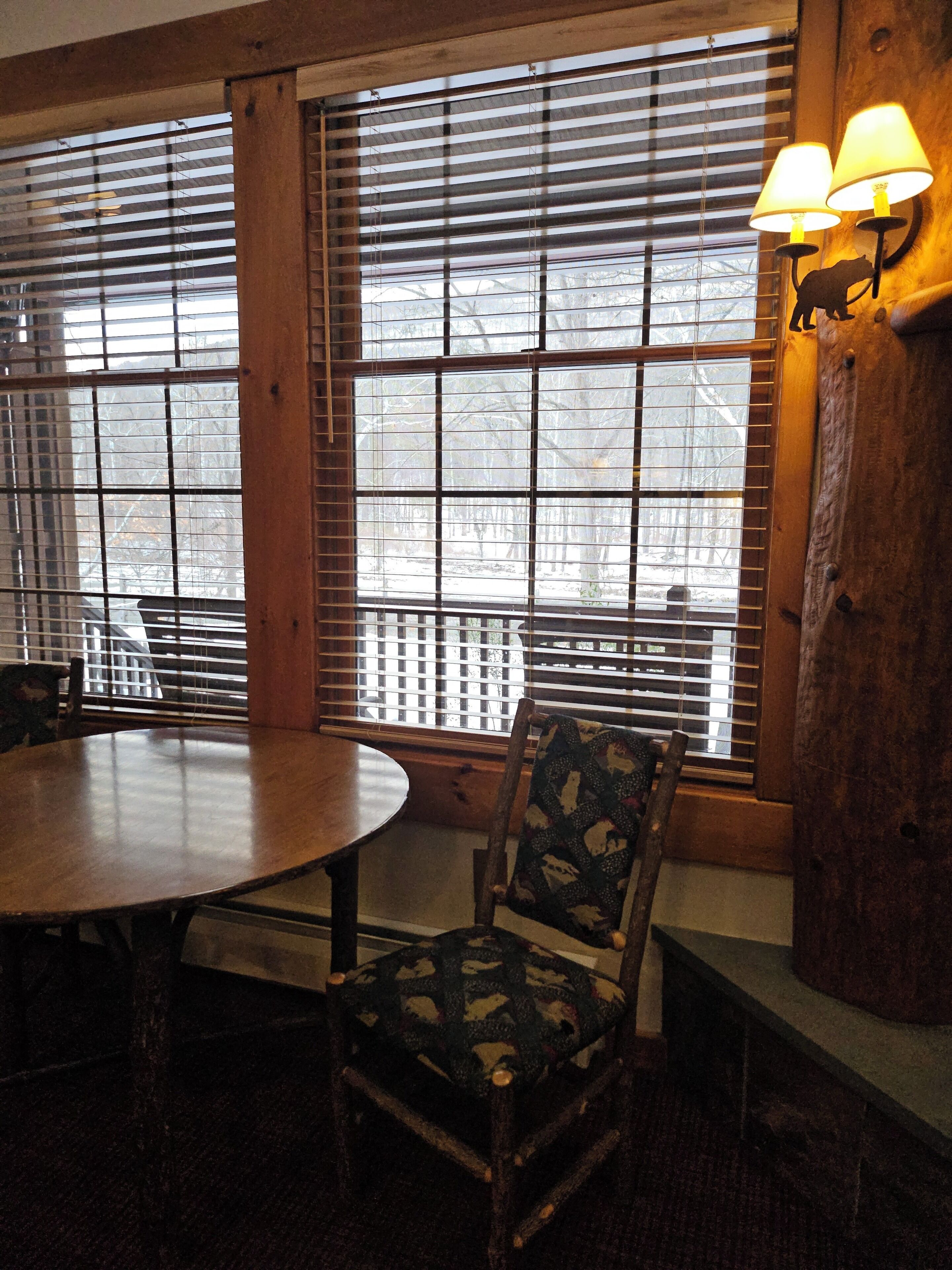 A room with a round wooden table and a chair with a patterned cushion. There are two windows with blinds, and a lamp is mounted on the wall.