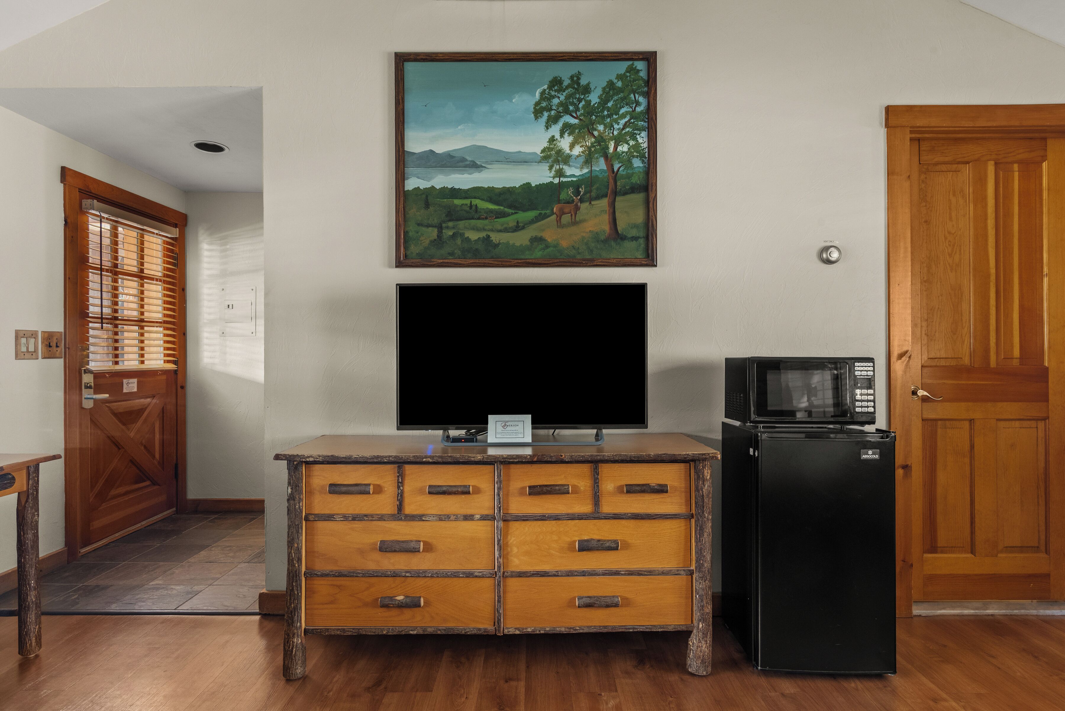 Una habitación con un armario de madera, un televisor de pantalla plana, un refrigerador negro y un cuadro de un paisaje en la pared.