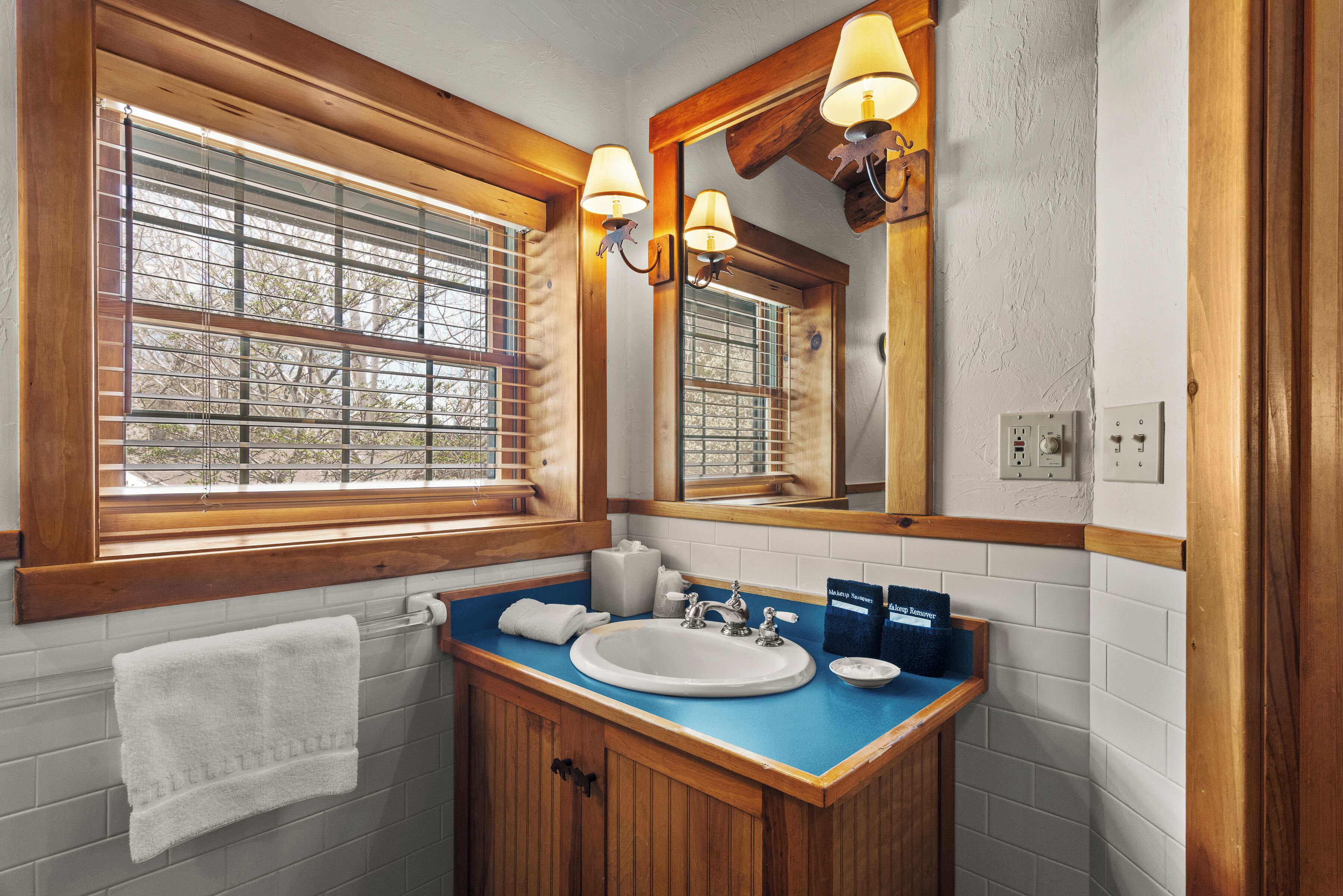A bathroom with a wooden vanity, a white sink, a mirror with wall sconces, and a window with blinds.