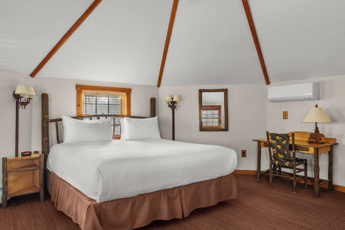 A bedroom with a large bed, wooden beams, a desk with a chair, and two wall-mounted lamps.