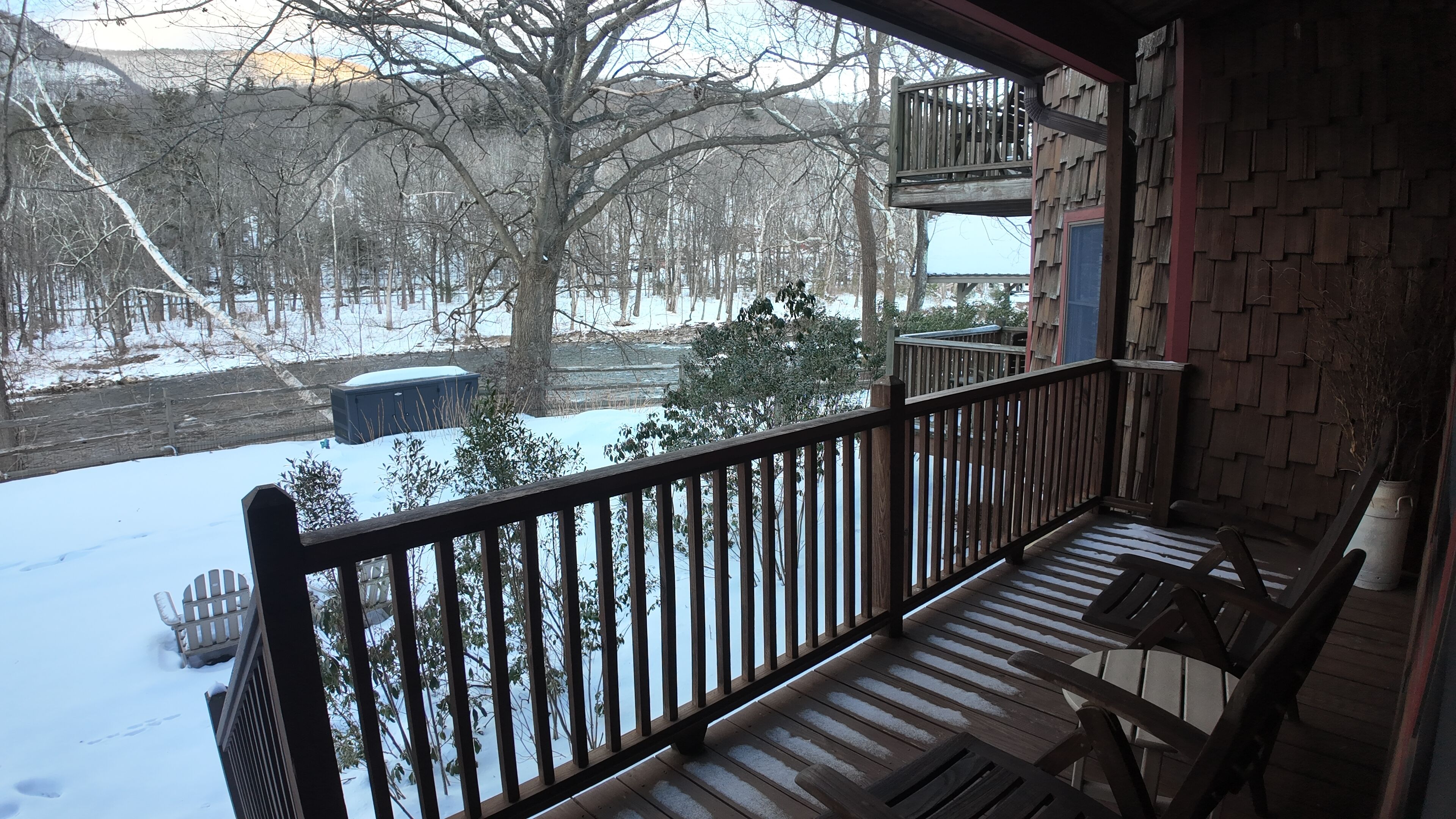 A wooden deck with a railing, snow-covered ground, and a view of a snowy landscape with trees and a building in the background.