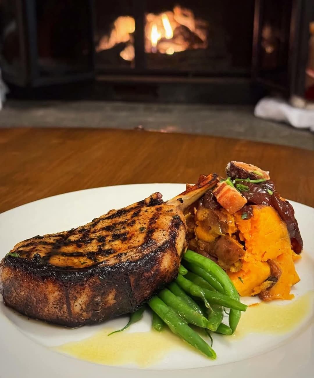 Une assiette de nourriture comprenant de la viande grillée, des haricots verts et un plat cuit au four, le tout posé sur une table devant une cheminée.