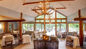 Un'ampia sala da pranzo con travi in legno, grandi finestre e un'isola centrale con tavoli e sedie.