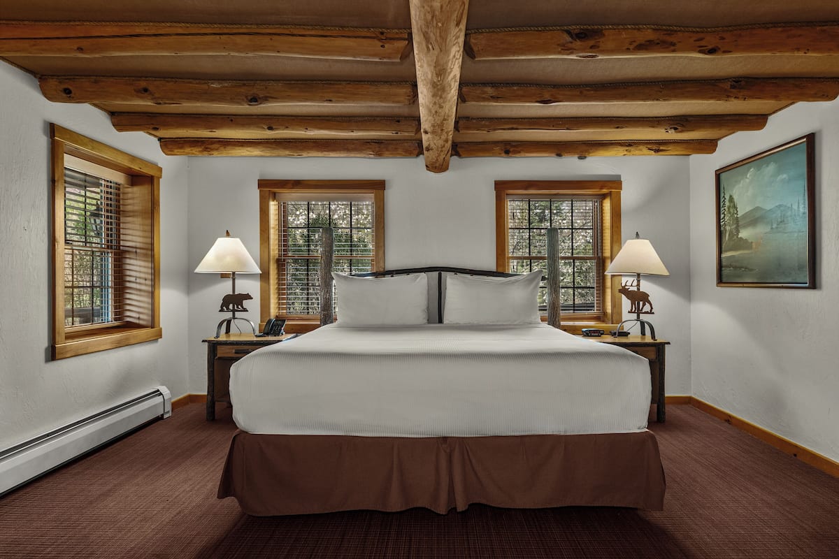 A bedroom with a large bed, two nightstands, a wooden ceiling, and windows with blinds.