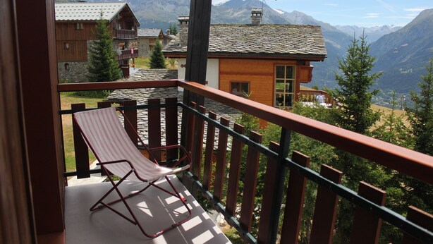 A balcony with a red chair, overlooking a mountain landscape and a wooden building with a sloped roof. - Studio quiet with stunning views over the valley (Montvalezan)