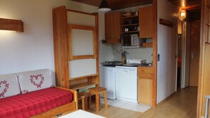A compact living space with a kitchenette, a sofa with a heart-patterned cushion, a small wooden table, and a white refrigerator. - Studio quiet with stunning views over the valley (Montvalezan)