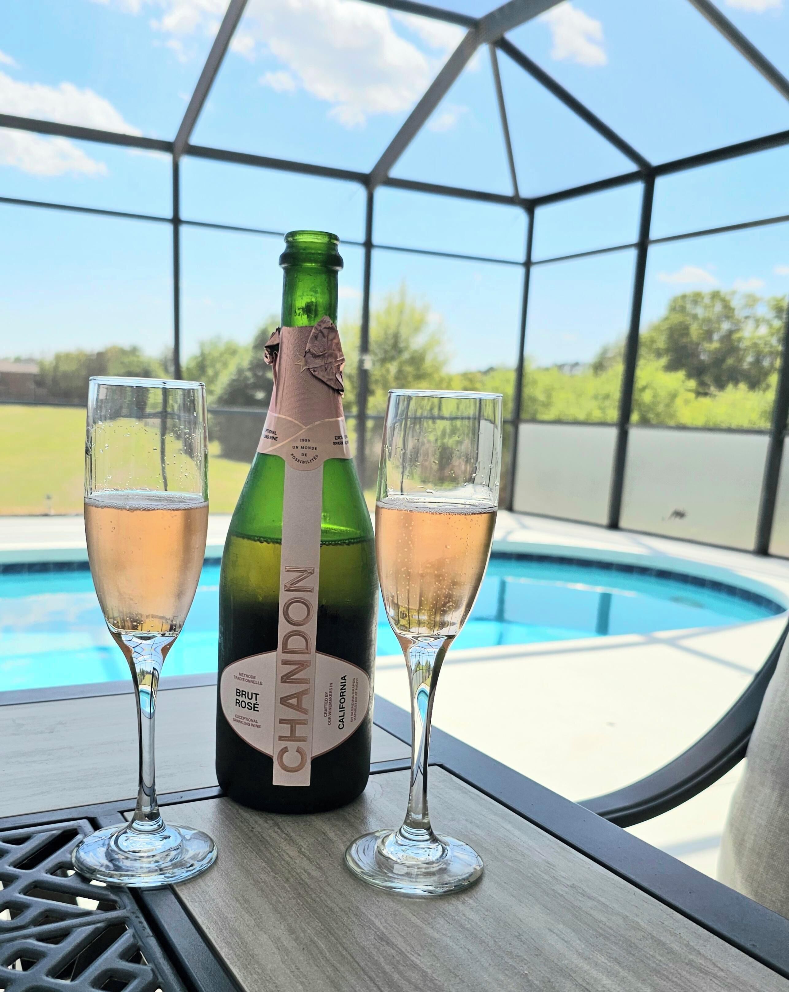 A bottle of Brut Rose wine and two glasses on a table by a pool.