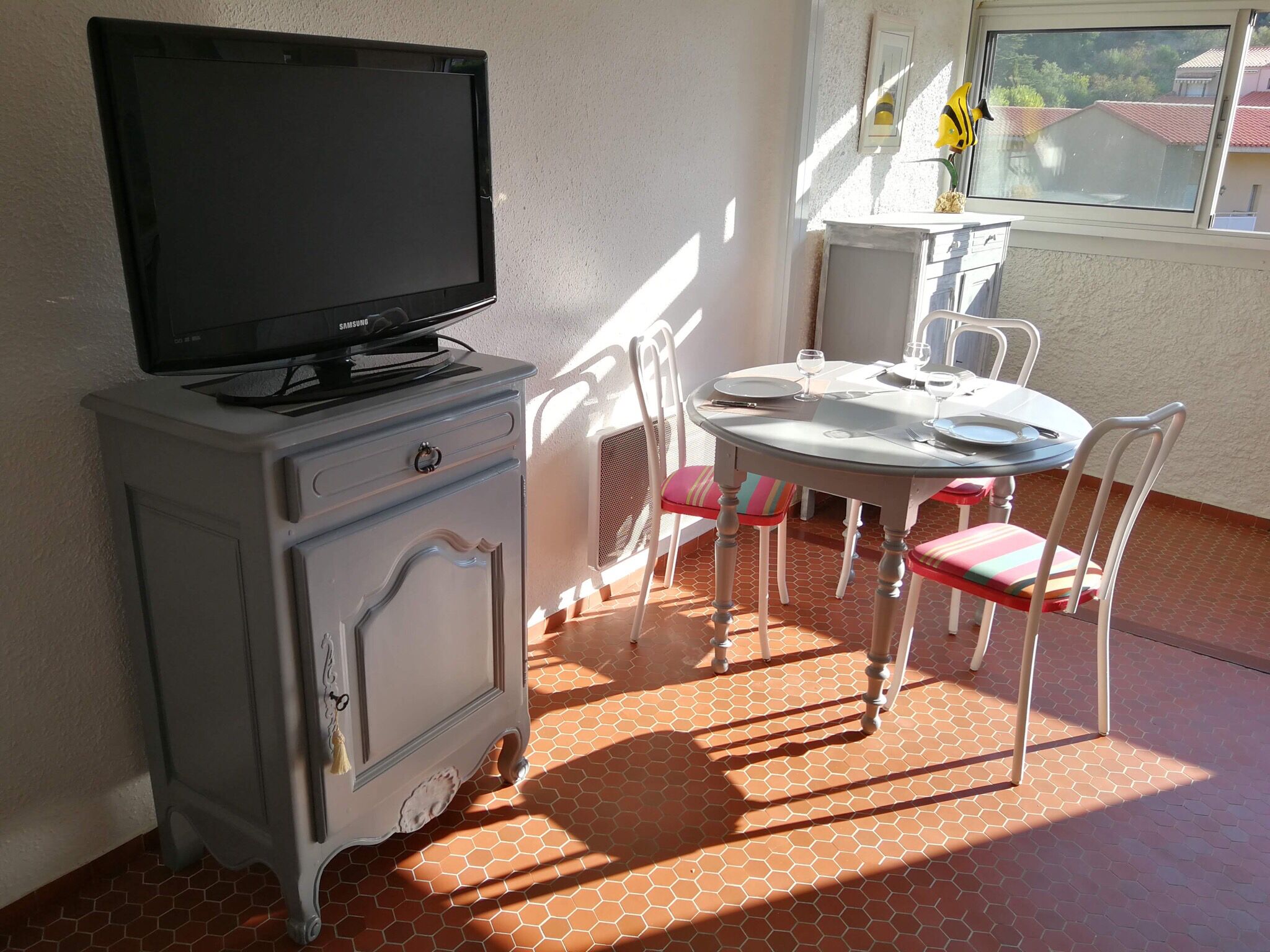 Une petite pièce avec un téléviseur posé sur un meuble, une table avec des chaises et une fenêtre donnant sur des immeubles.