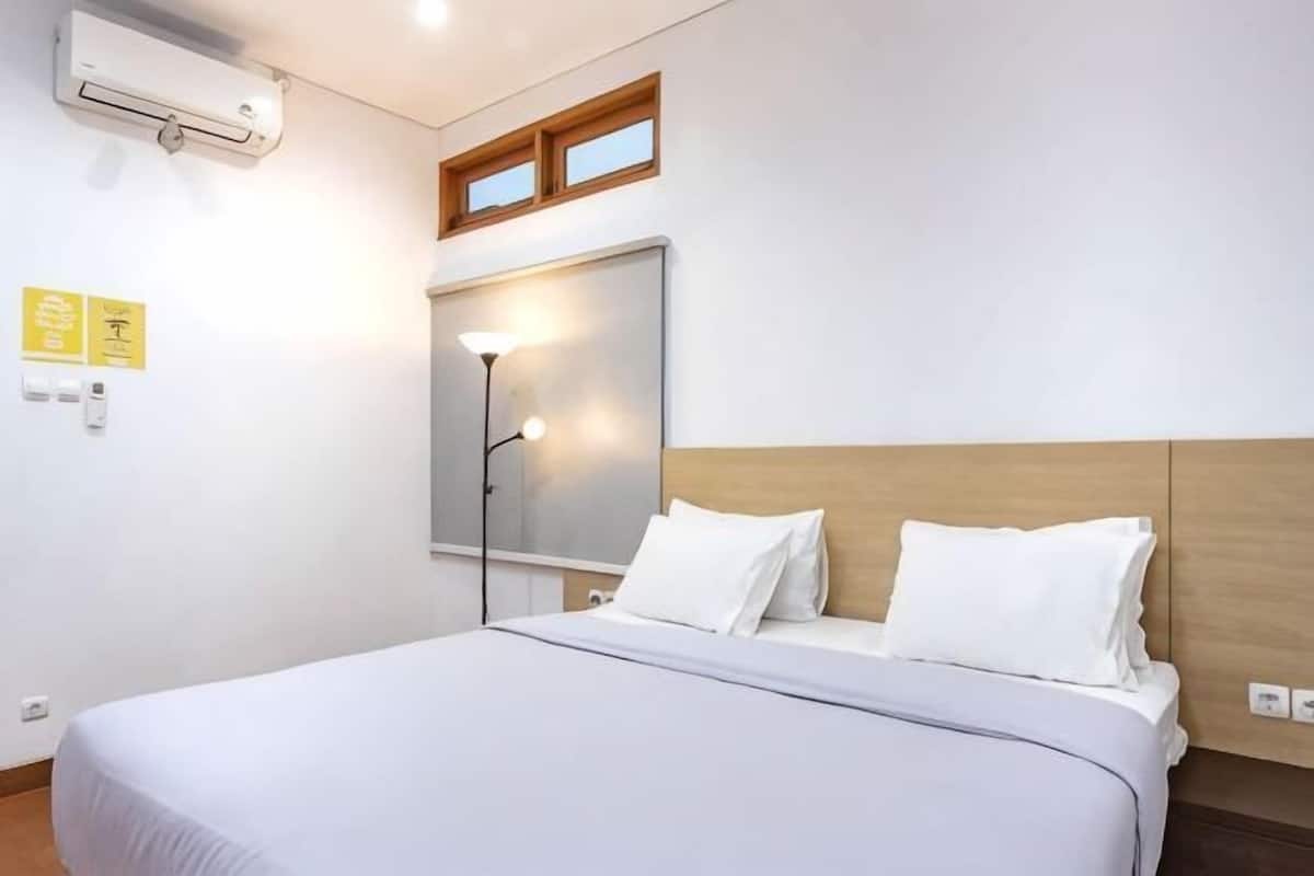 A hotel room with a bed, a nightstand, a wall-mounted air conditioner, a window, and a wall-mounted light fixture.