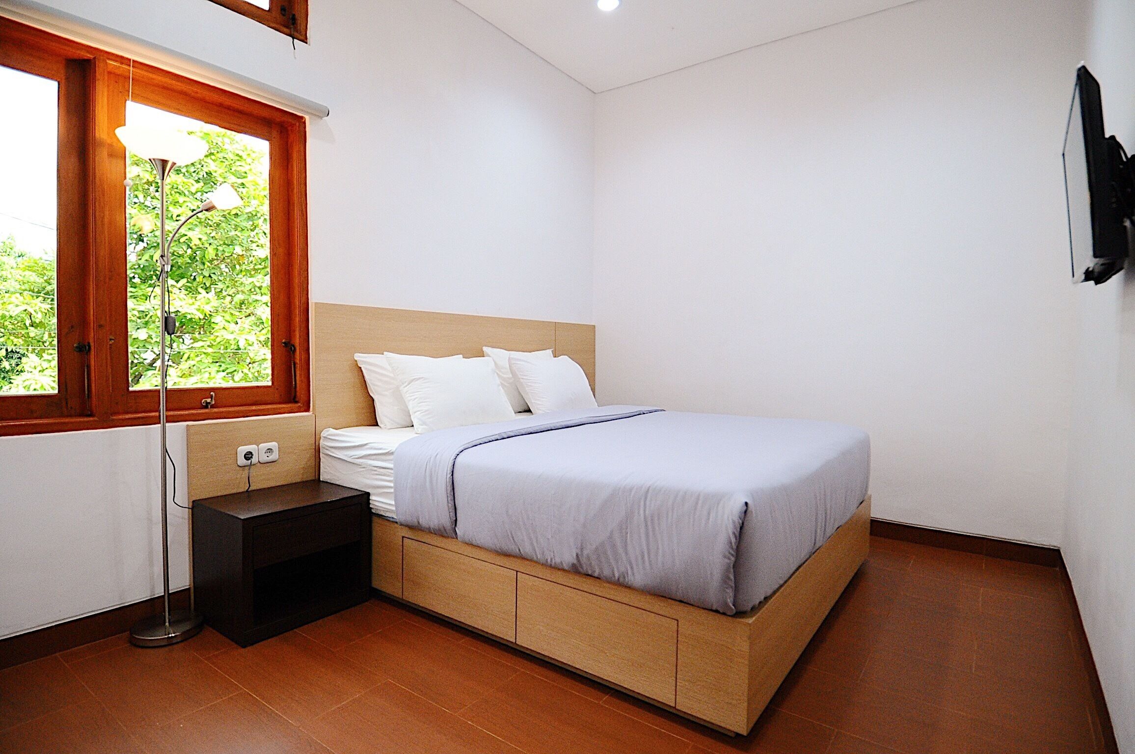 Quarto com cama, mesa de cabeceira, abajur e janela com vista para o verde.