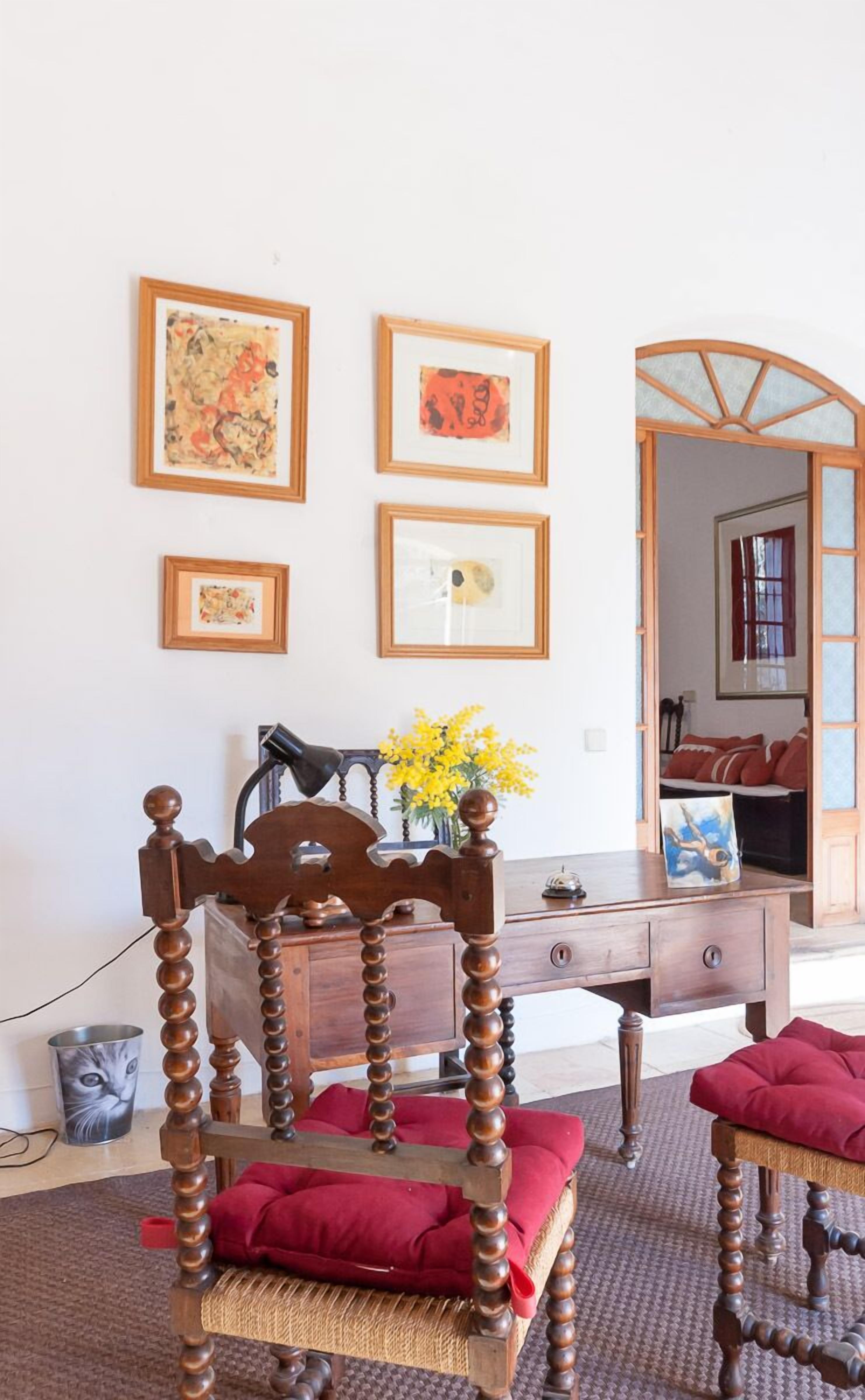 A room with a wooden chair, a desk, and a mirror. There are framed pictures on the wall and a vase with yellow flowers on the desk.