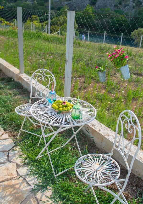 Een metalen tafel met stoelen, een mand met fruit, en een glas met rietje, op een stenen terras met een gazon en een hek op de achtergrond.