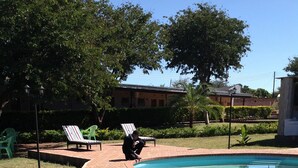A pool area with a person sitting on a chair, surrounded by grass and a brick border. - Eland Lodge (Livingstone)