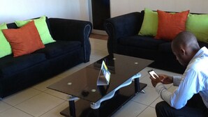 A room with two dark-colored sofas with bright green and orange cushions, a glass-top coffee table, and a person using a smartphone. - Eland Lodge (Livingstone)