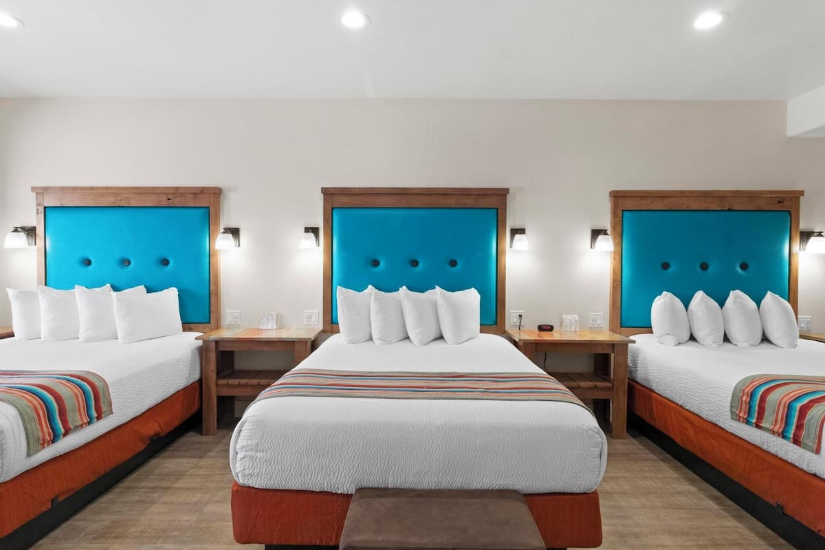 Een hotelkamer met drie bedden, elk voorzien van een houten hoofdbord en wit beddengoed. Er zijn nachtkastjes met lampen en een bankje tussen de bedden.