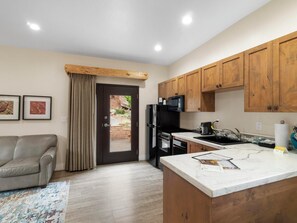 A modern kitchen with wooden cabinets, a black refrigerator, a stove, and a microwave. There is a gray sofa and a patterned rug in the living area.