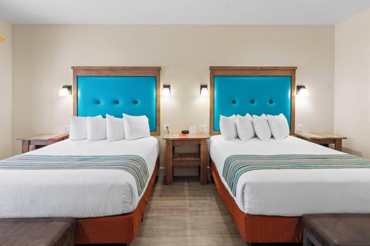 Een hotelkamer met twee bedden, elk met een houten hoofdbord en witte beddengoed. Er staan nachtkastjes met lampen en een klein tafeltje tussen de bedden.