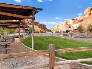 A rustic outdoor area with picnic tables and a wooden pergola, surrounded by a gravel path and a well-maintained lawn.