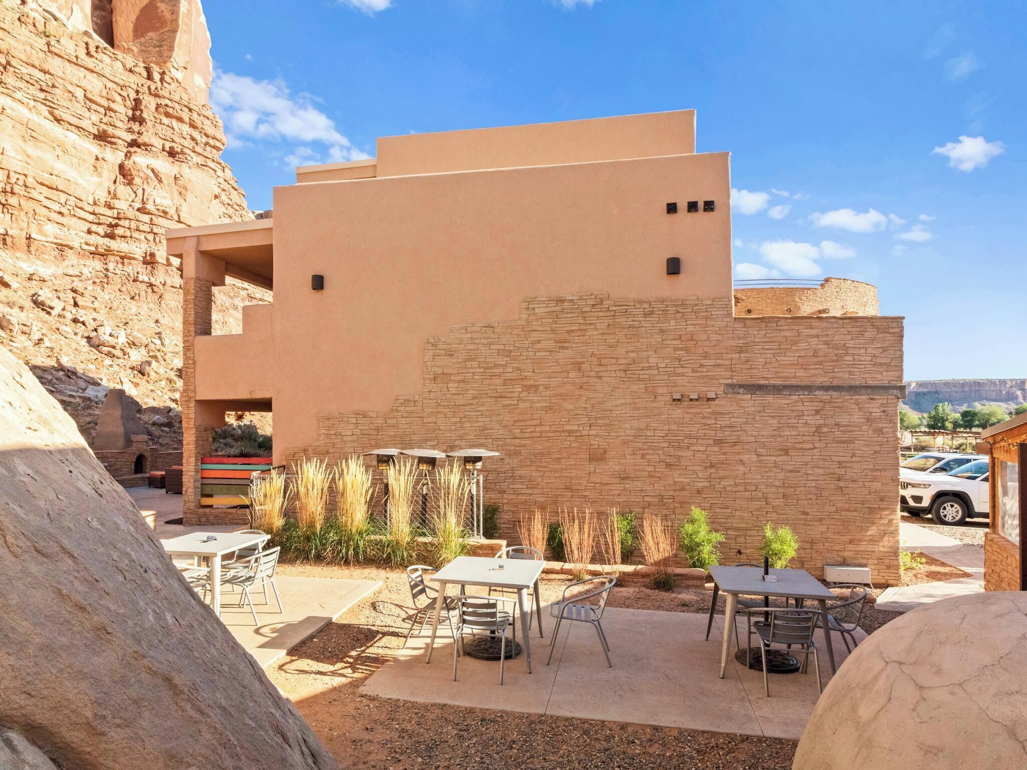 Un bâtiment moderne doté d’un toit plat et d’une façade en pierre, avec un espace extérieur aménagé avec des tables et des chaises, et un ciel dégagé.