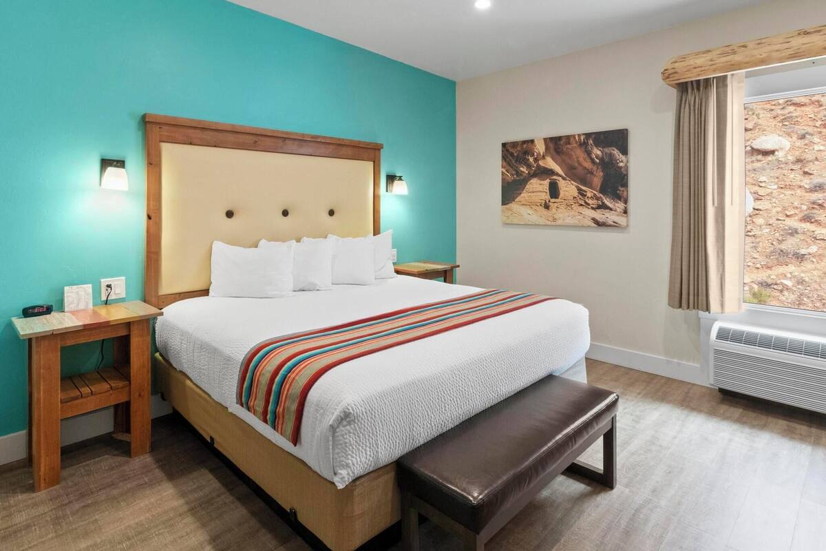 Een slaapkamer met een bed, een houten nachtkastje, een bankje en een schilderij aan de muur.