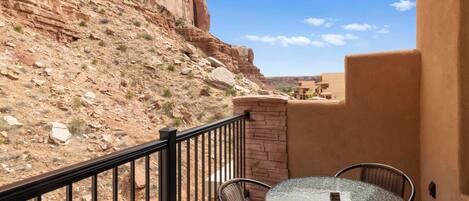 A balcony with a round glass table and wicker chairs, overlooking a rocky landscape and a building.