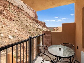 Ein Balkon mit rundem Glastisch und Korbstühlen, mit Blick auf eine felsige Landschaft und ein Gebäude.
