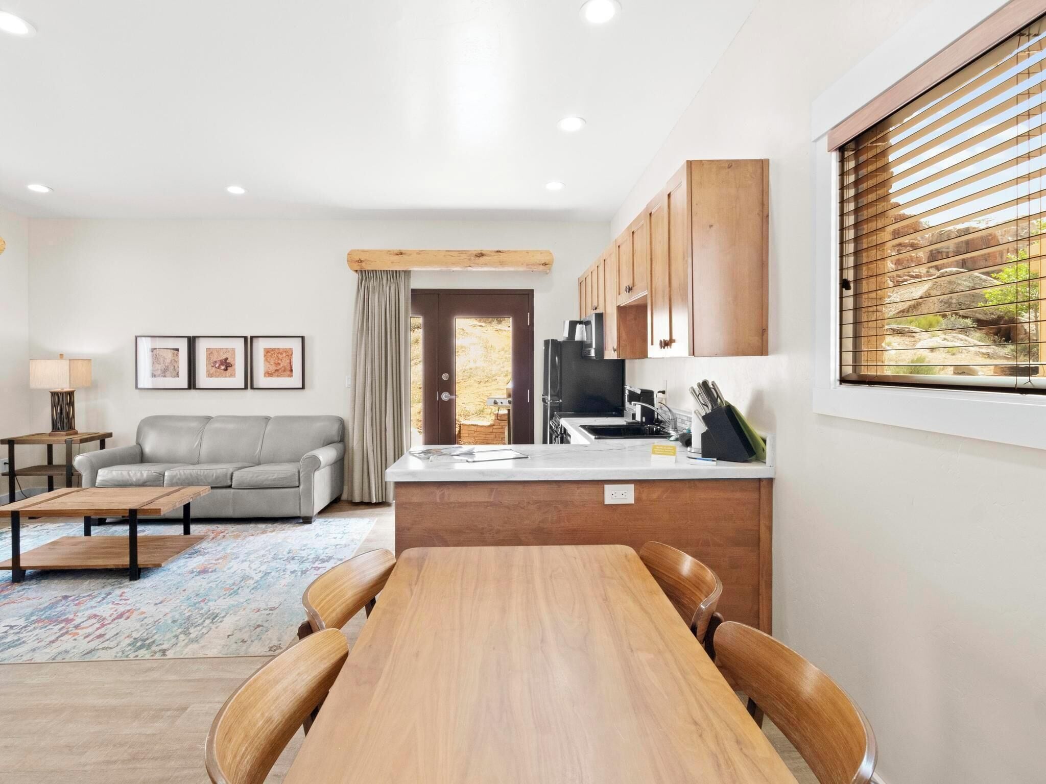 A modern living room with a sofa, a dining table with chairs, a kitchen area with wooden cabinets, and a window with blinds.