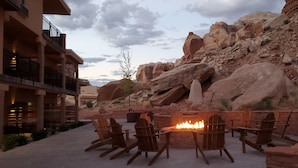 A patio with wooden chairs and a fire pit, surrounded by large rocks and a building with balconies.