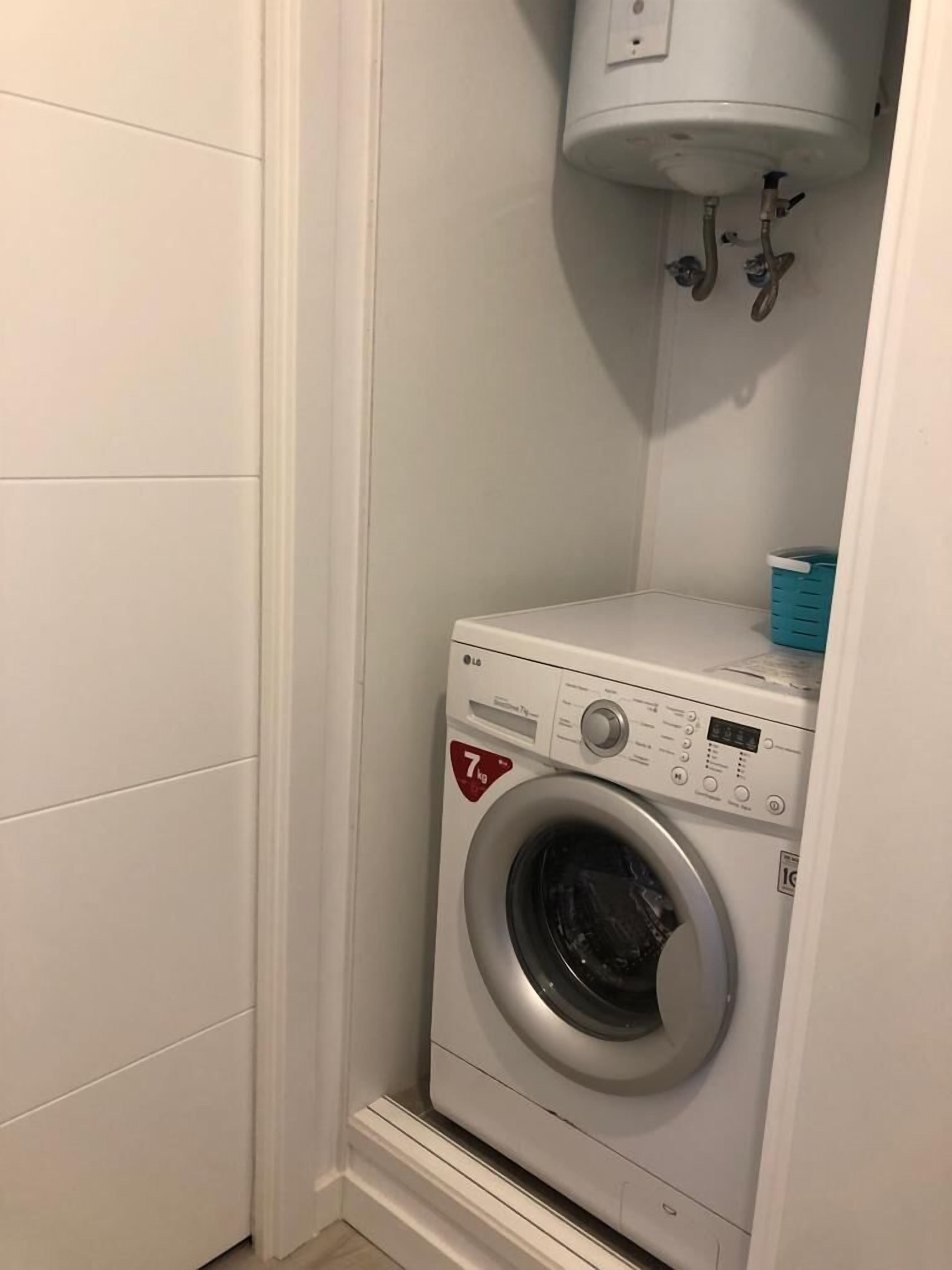 Une petite buanderie équipée d’un lave-linge à chargement frontal, d’un petit panier à linge bleu et d’un chauffe-eau blanc fixé au-dessus de la machine.