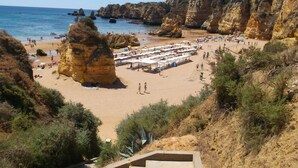 Ein Strand mit felsigen Klippen, einem Sandbereich mit Sonnenschirmen und einer Treppe, die hinunterführt.