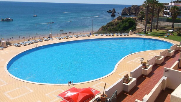Ein großer, gebogener Pool mit Blick auf das Meer und Boote. Am Poolufer stehen Liegestühle und Sonnenschirme.