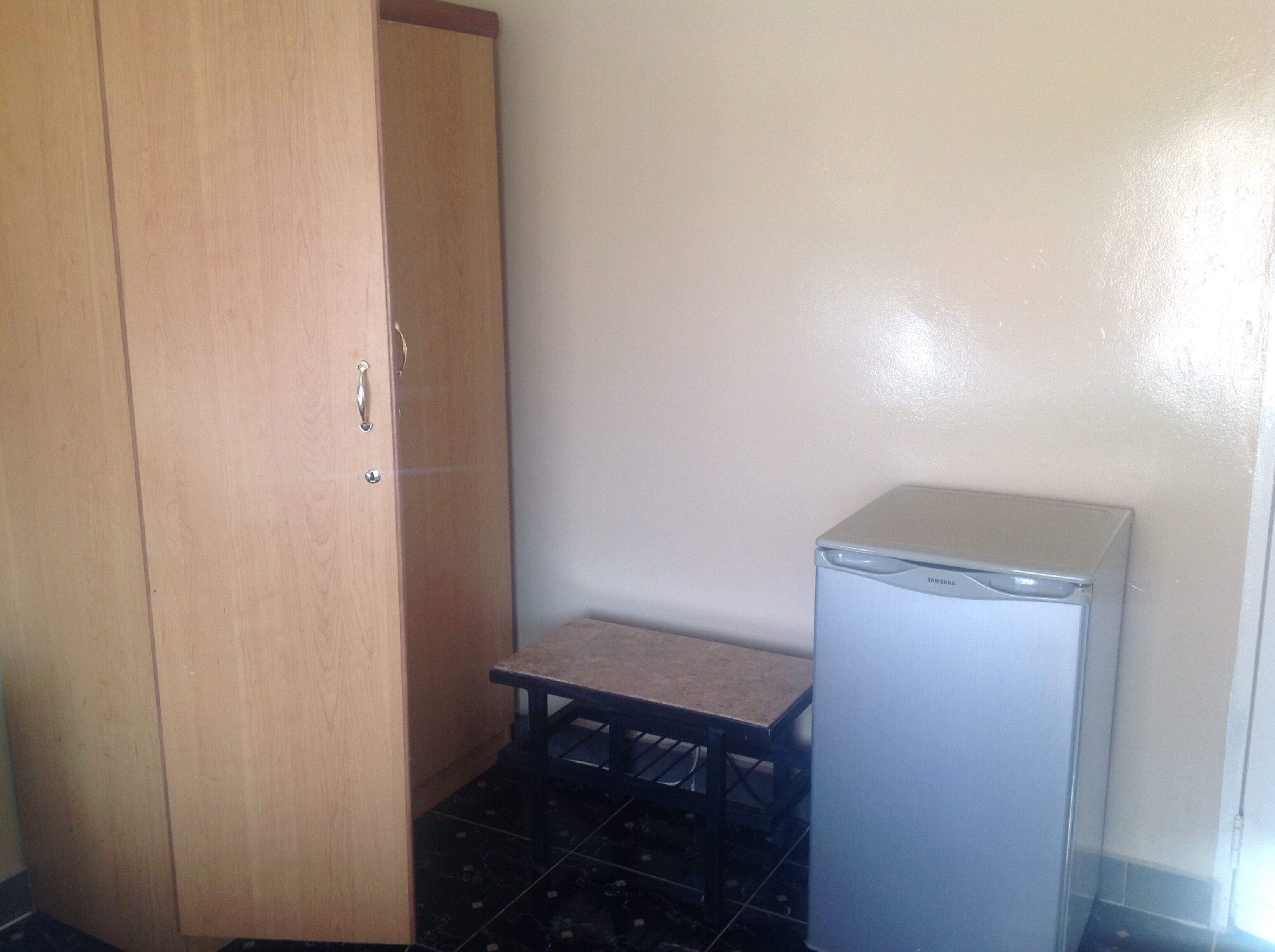 A small room with a wooden wardrobe, a small table, and a refrigerator.