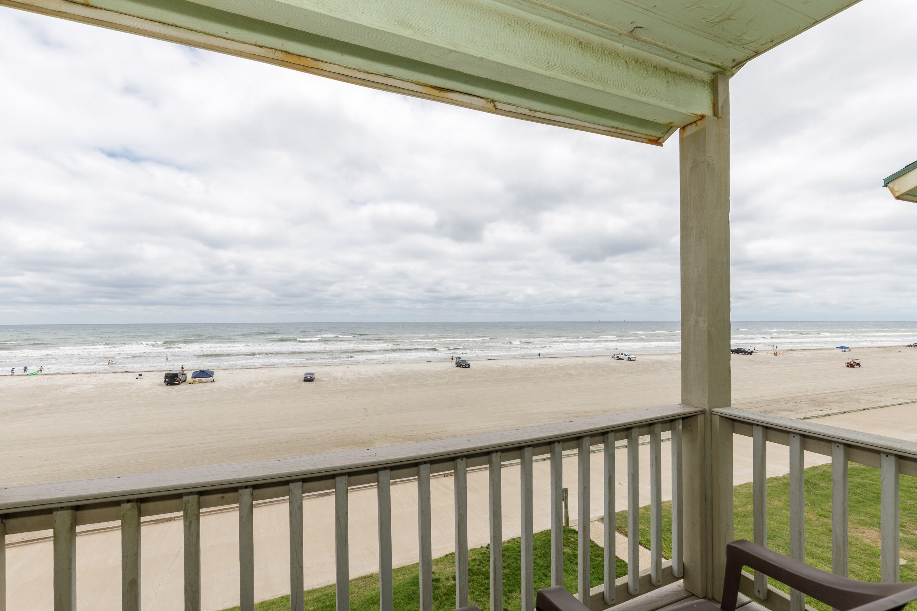 A beach view from a covered balcony with a railing, a bench, and a grassy area.