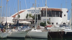 Un port de plaisance où sont amarrés plusieurs bateaux, un bâtiment portant l’inscription « RESTAURANTE NAUTICO » et coiffé d’un toit en tuiles, ainsi qu’un drapeau flottant sur le bâtiment.