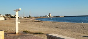 A beach with a paved walkway, a lighthouse, and sailboats in the background.