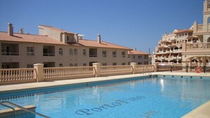 Une piscine à l’eau d’un bleu limpide, entourée d’une clôture beige et, au fond, un bâtiment doté de balcons.