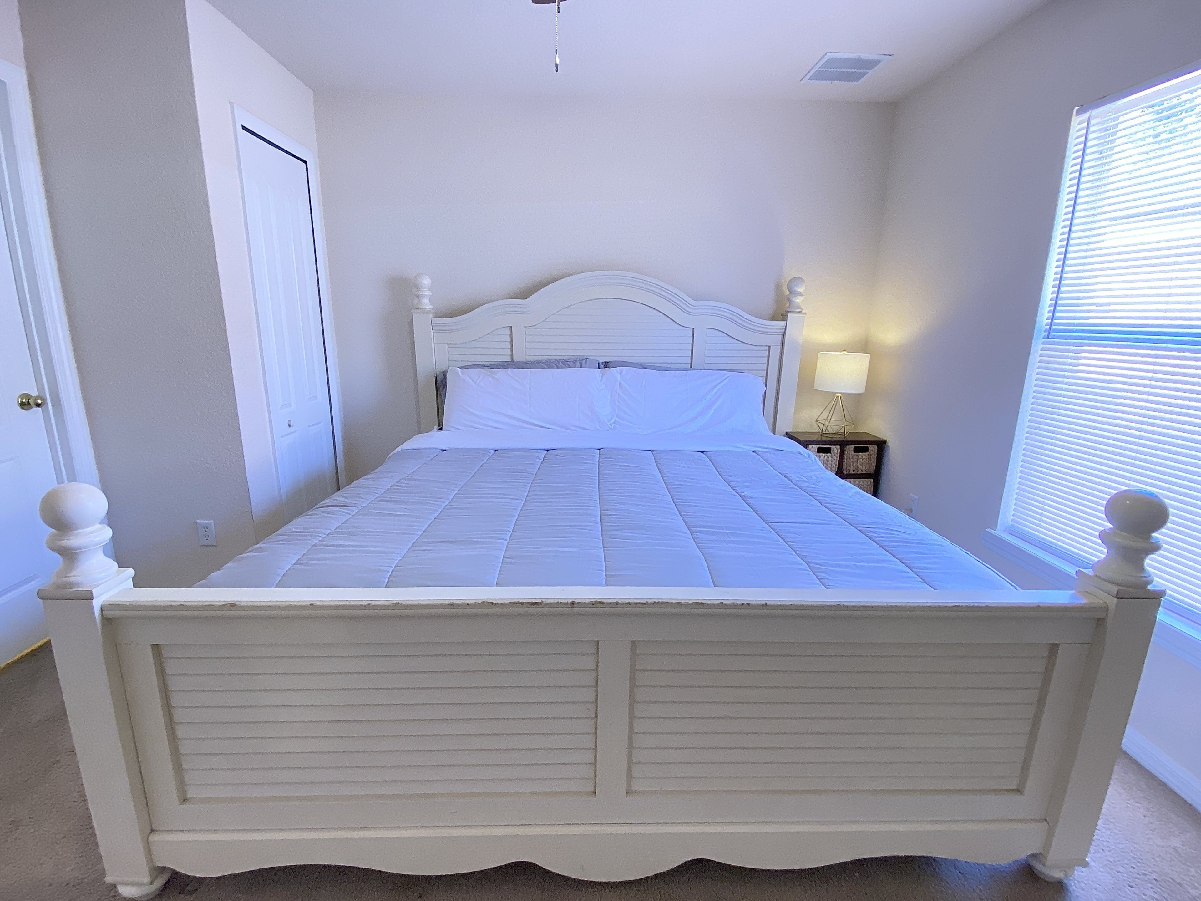 A white bed with a headboard, a bedside table with a lamp, and a window with blinds.
