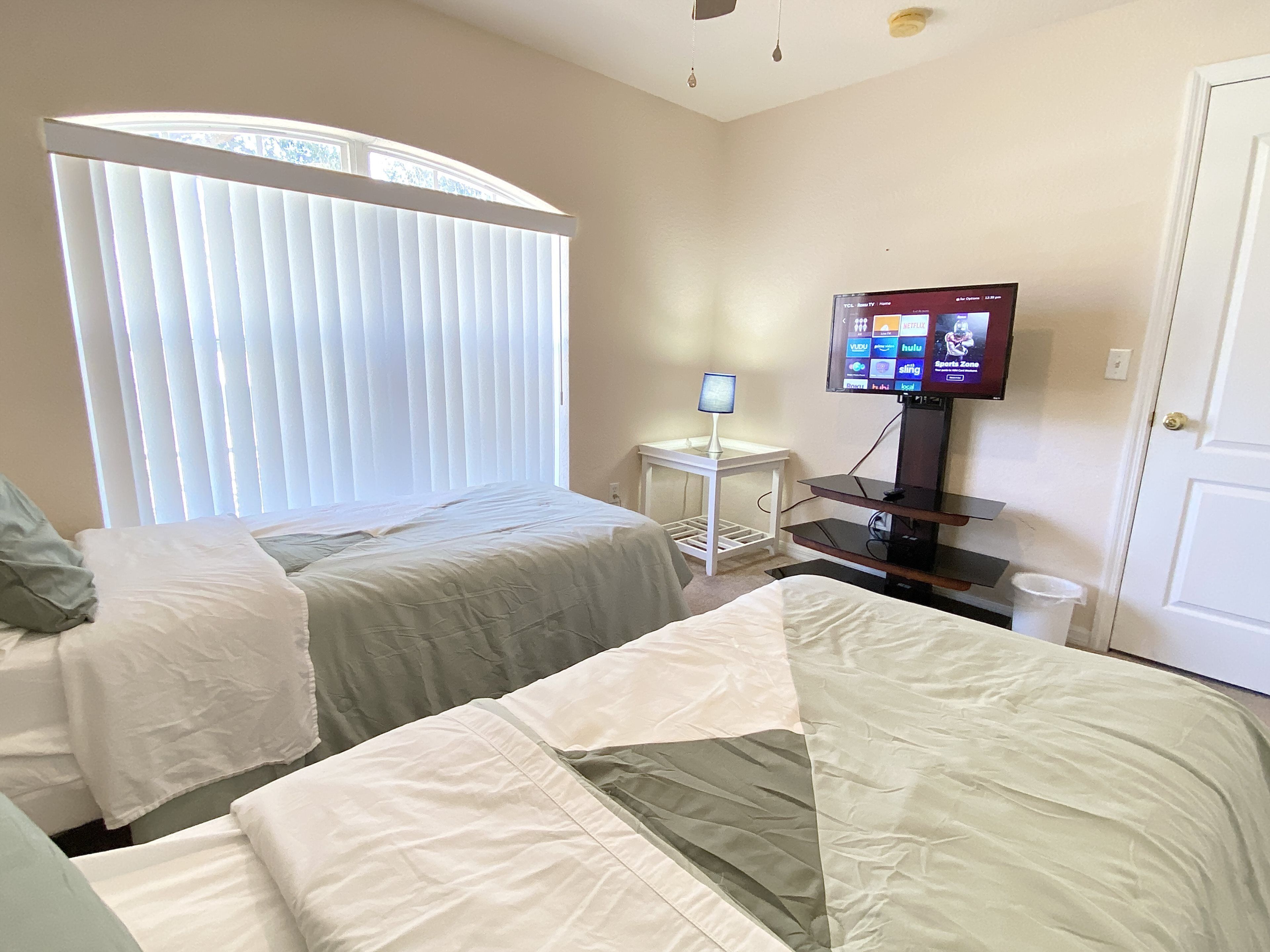 A hotel room with two beds, a TV on a stand, a small table with a lamp, and a window with blinds.
