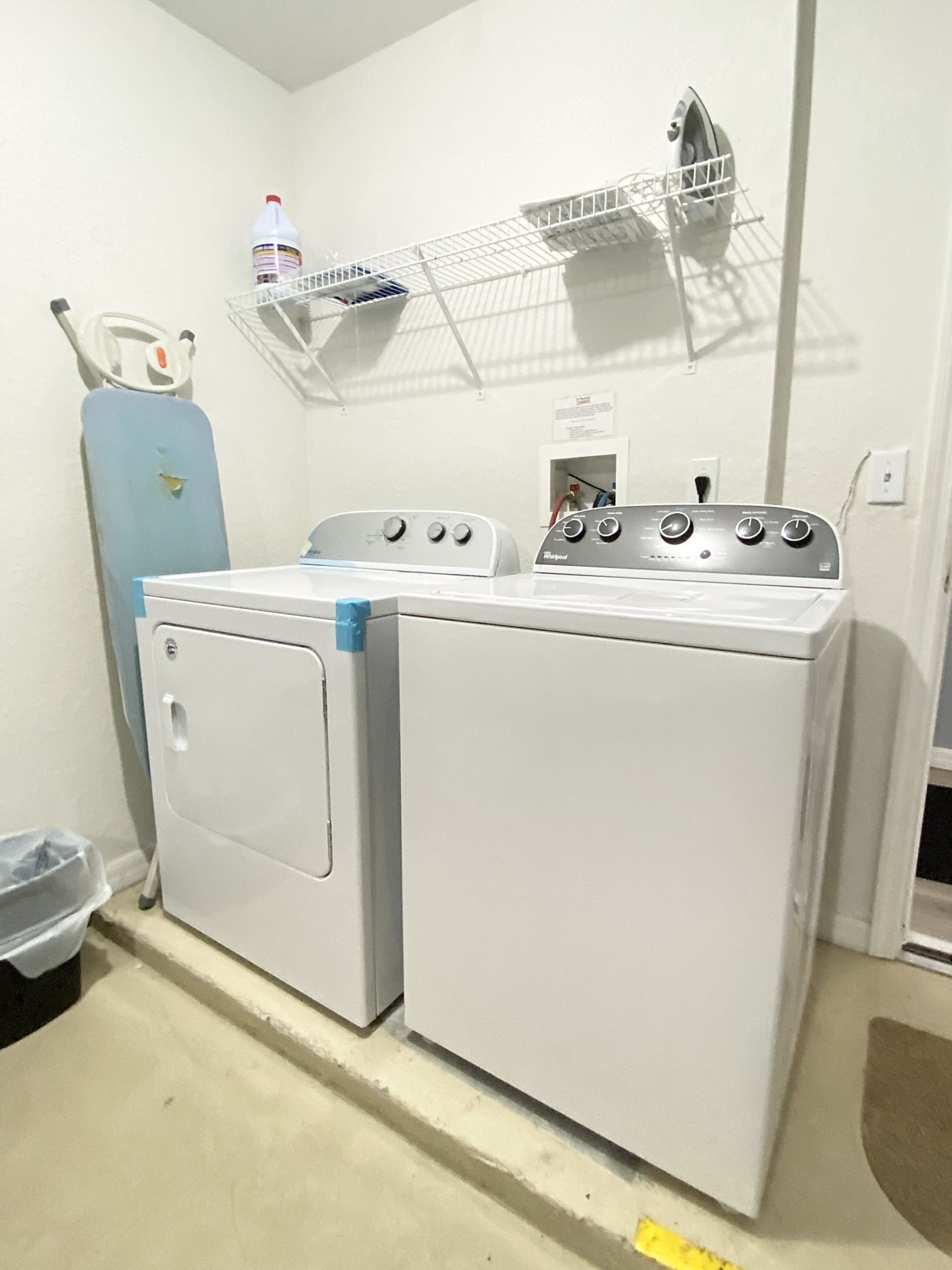 A laundry area with a front-loading washer and dryer, a blue water heater, and a metal shelf above the dryer.