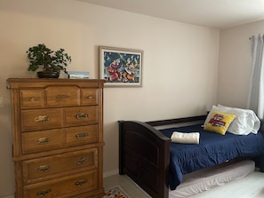 A bedroom with a wooden dresser, a bed with blue bedding, a painting on the wall, and a window with a curtain.