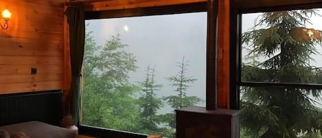 A cozy cabin interior with a large window, a sofa, a wooden ceiling, and a fireplace.