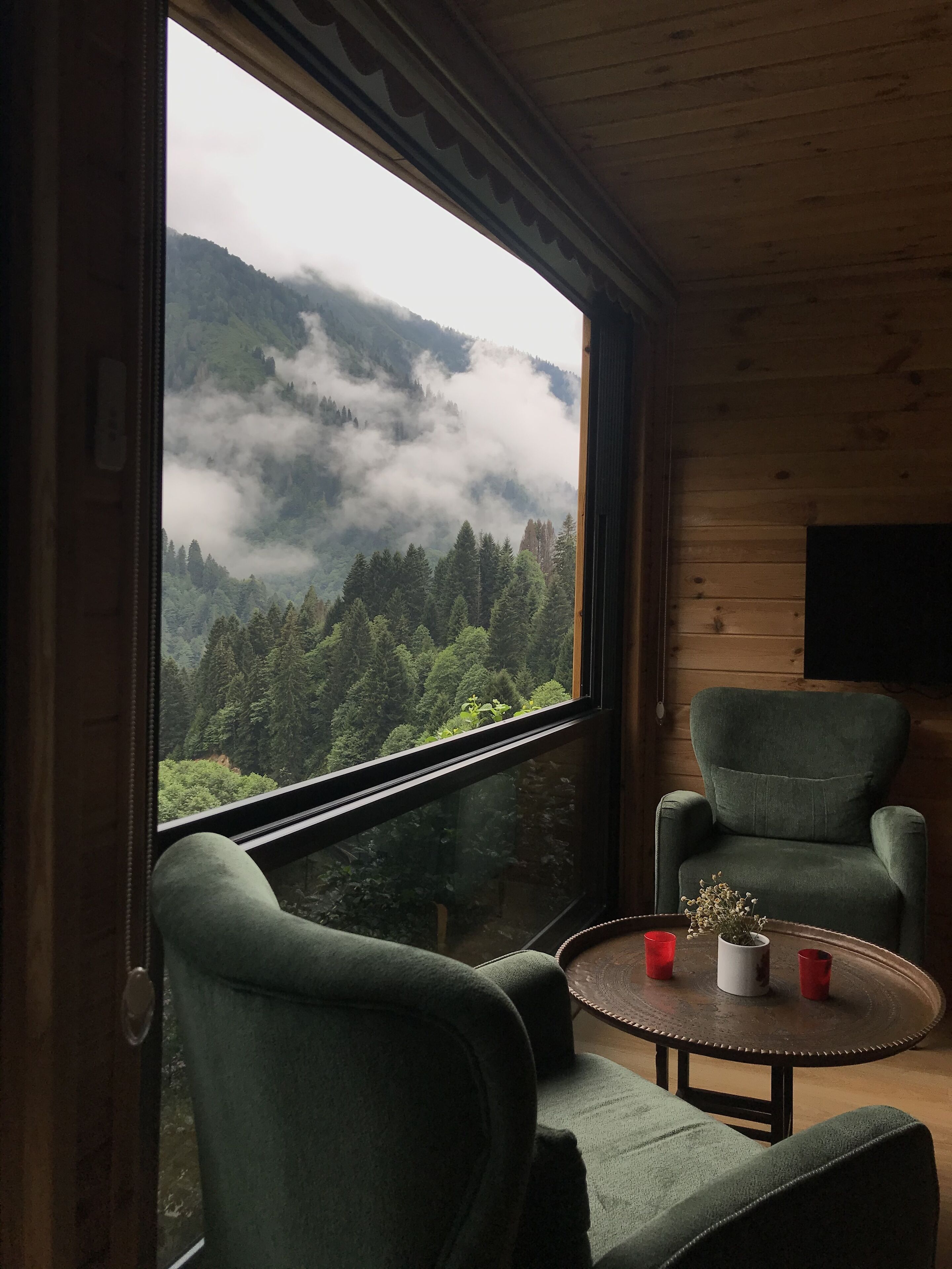 Una habitación con un ventanal que ofrece vistas a una cadena montañosa boscosa y nubes. En el interior, hay una mesa redonda con dos sillas y una pequeña planta en maceta.