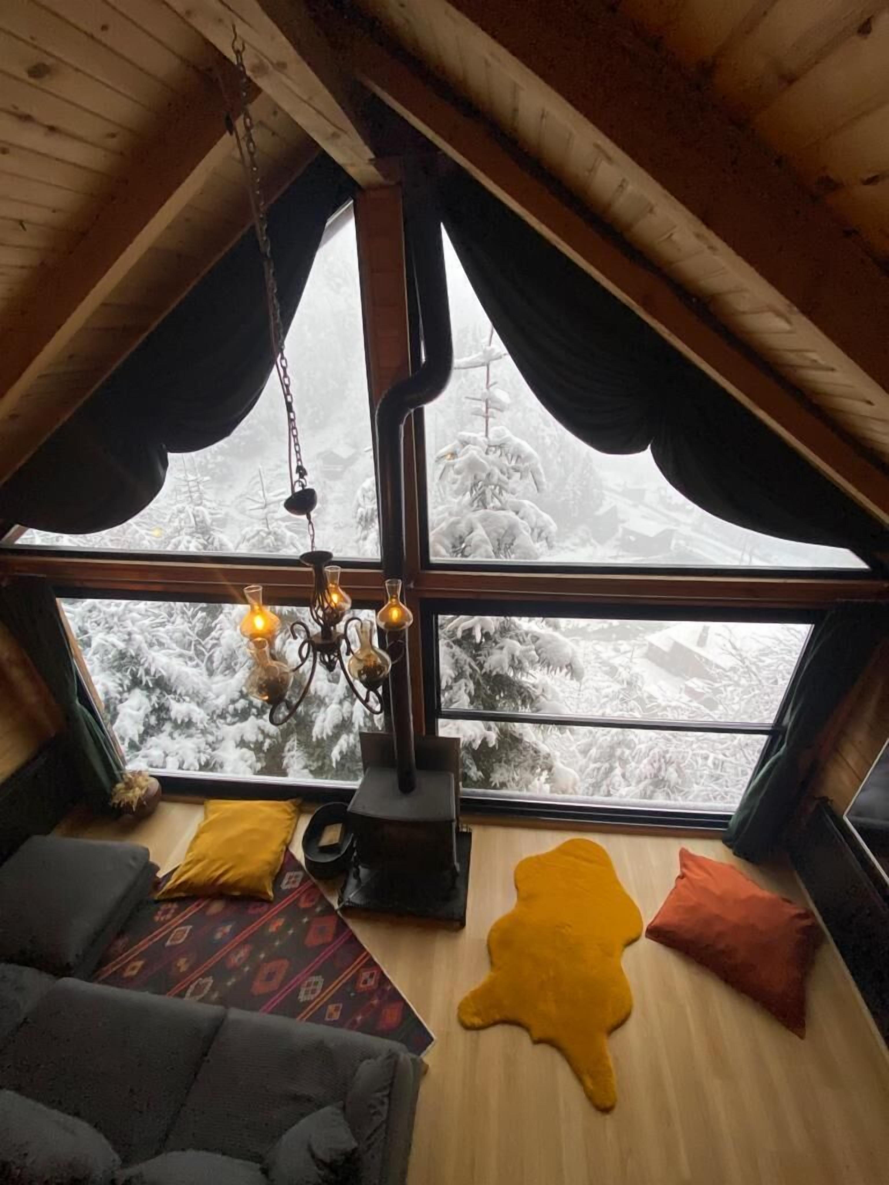 A cozy room with a high ceiling, a fireplace, and a large window offering a snowy view.