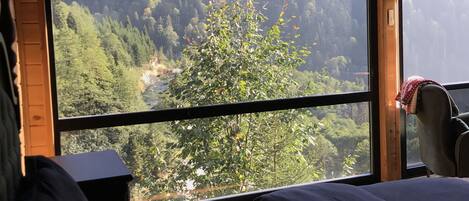 A room with a bed, a chair, and a large window offering a view of a forested mountain area.