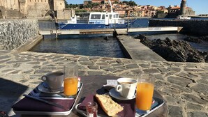 A table with breakfast items including coffee, orange juice, and a pastry, overlooking a harbor with boats and historic buildings. - House in beautiful Collioure with pool and sea views from both balconies (Collioure)