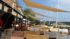 An outdoor dining area with tables and chairs under large canopies, overlooking a rocky hillside.