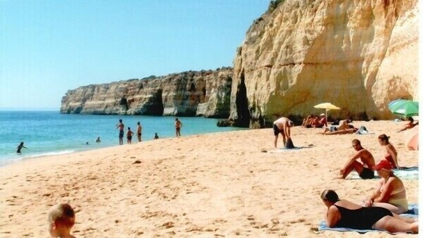 A beach with people sunbathing and swimming, a cliff in the background, and umbrellas on the sand.